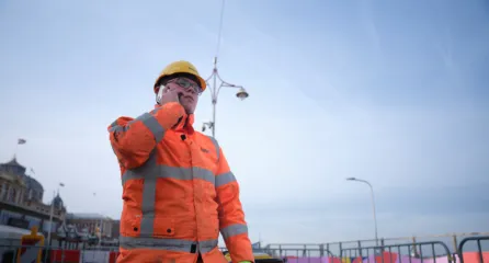 Profielfoto bij de functie werkvoorbereider. Deze man met helm en veiligheidskleding voert een telefoongesprek op de bouwplaats.