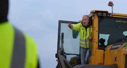 Profielfoto van uitvoerder sector infra. Bouwplaatsmedewerker in gele veiligheidsjas geeft aanwijzingen vanuit de cabine van een shovel.