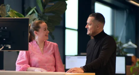Profielfoto bij de functie manager. Twee collega’s staan lachend bij een bureau op kantoor en praten met elkaar.