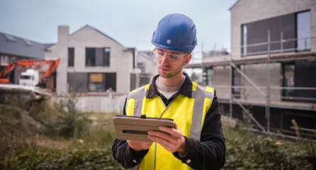 Profielfoto van een veiligheidskundige met blauwe helm en Ipad in zijn hand voor twee huizen in aanbouw