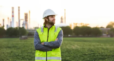 Profielfoto van functie diagnose specialist. Man met geel veiligheids hesje en witte helm voor een groen veld met een fabriek in de achtergrond.
