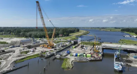 Profielfoto van brug over een rivier in aanbouw met een grote gele hijskraan en een binnenvaartschip