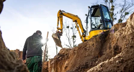 Profielfoto bij functie vakman grond water weg in de sector infa. Een vakman staat in een greppel met schep in zijn hand met een kleine gele graafmachine in de achtergrond.