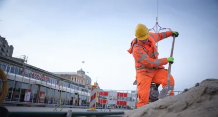 Profielfoto van de functie van bouwplaatshelper sector Infra. Bouwplaatshelper verplaatst stalen balken met een kraan tussen twee vrachtwagens op het strand.