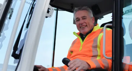 Profielfoto bij functie machinist sector infra. Machinist in oranje veiligheidskleding glimlacht achter het stuur van een graafmachine.