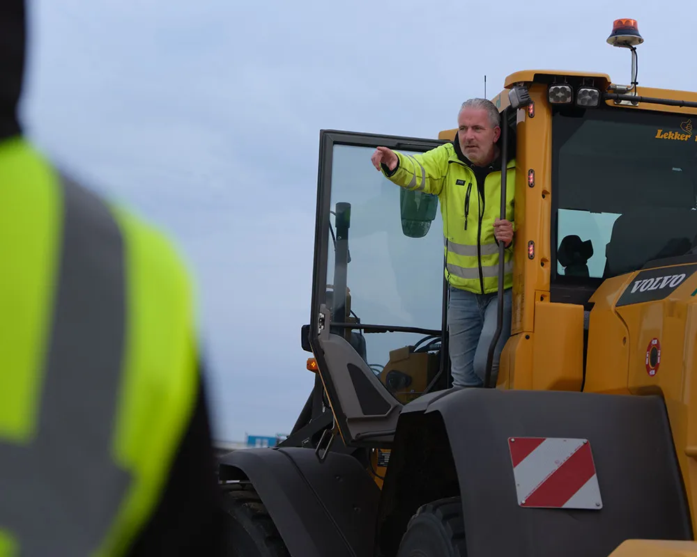 Profielfoto van uitvoerder. Man in gele veiligheidsjas geeft aanwijzingen vanuit de cabine van een shovel.