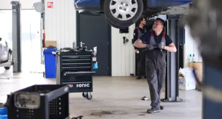 Profielfoto bij de functie monteur in de sector mobiliteit. Man loopt op werkplaats onder auto door die opgehangen is om de auto te inspecteren.