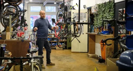 Profielfoto bij de functie eerste fietstechnicus in de sector mobiliteit. Man praat met collega buiten beeld en staat naast een fietswerkstandaard in een werkplaats vol met fietsen en onderdelen.