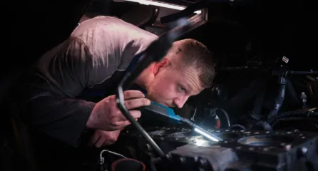 Profielfoto bij de functie eerste monteur in de sector mobiliteit. Werknemer onderzoekt de motor van een auto met een lampje.